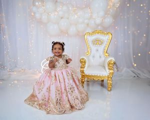 Baby girl in pink gold gown stands beside ornate white and gold throne with white balloon backdrop.