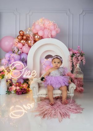 Baby girl in purple tutu sits on white chair with rainbow arch, balloons, and One neon sign for first birthday.