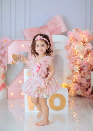 Toddler girl in pink ruffled dress stands on illuminated TWO letters with bow and floral backdrop.