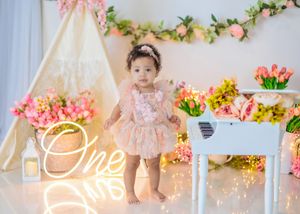Baby girl in pink floral dress stands near white piano with tulips, teepee, and glowing One neon sign.