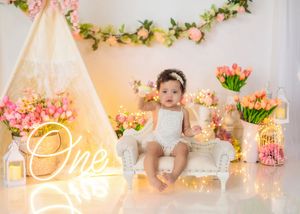 Baby girl in white outfit sits on small chair near teepee with tulips and glowing One neon sign.