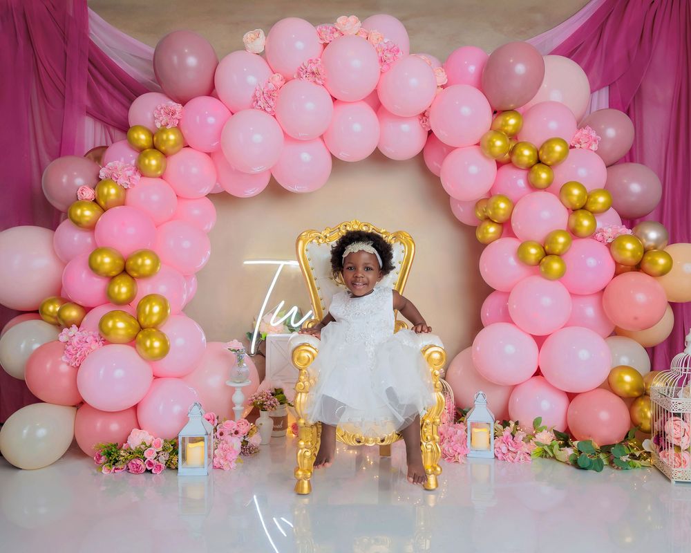 Young girl in white dress sits on gold throne chair at pink and gold balloon arch first birthday celebration.