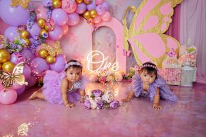 Twin baby girls in purple outfits pose before pink purple balloon arch with neon 'One' sign.