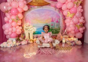 Toddler girl in pink dress celebrates first birthday with princess castle balloon arch backdrop.