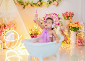 Baby girl in purple outfit sits in white mini bathtub wearing floral crown surrounded by tulips and lights.