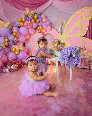 Two baby girls in purple tutus pose with balloon arch, butterfly decor, and floral arrangements on pink backdrop.