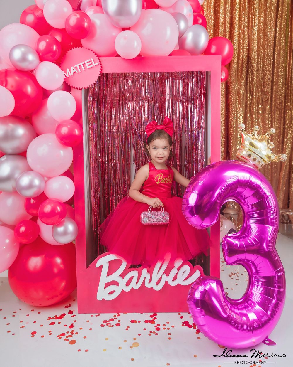 Young girl in red ball gown poses inside Barbie box frame at pink and gold third birthday party.