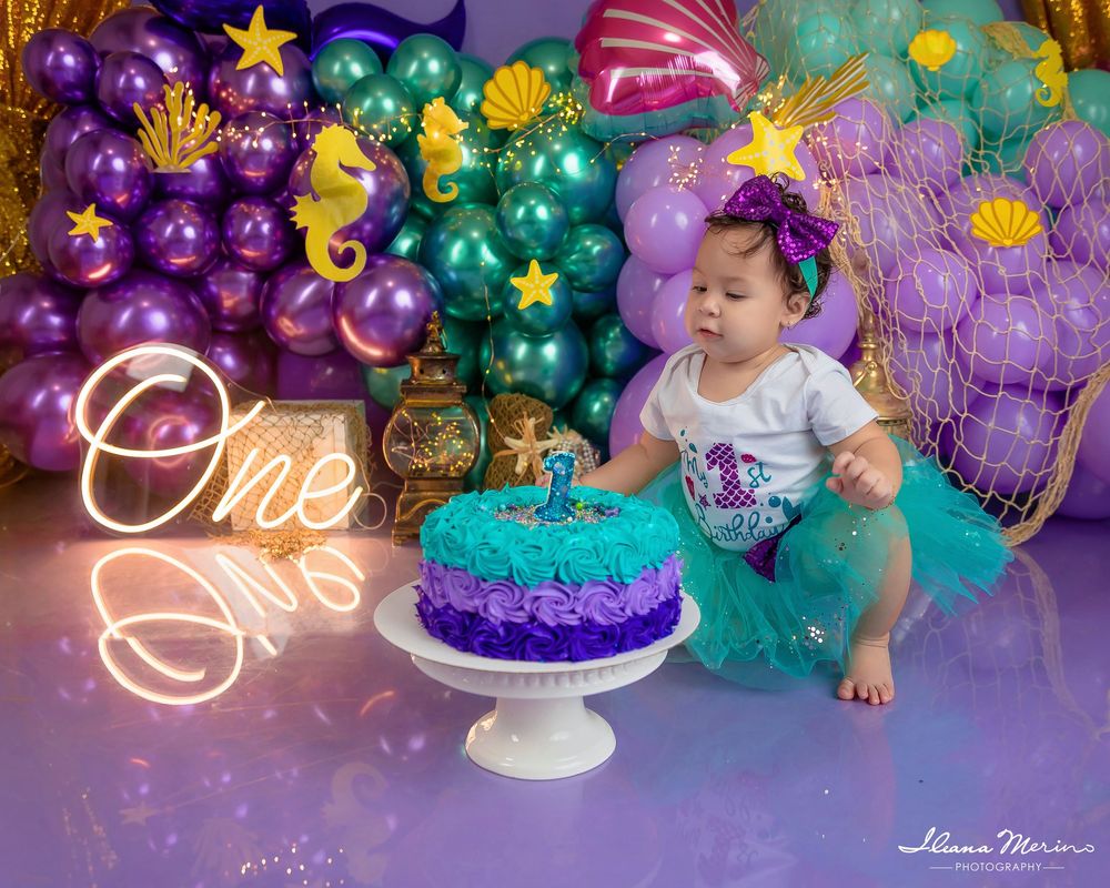 Baby girl in teal tutu sits with mermaid cake at Little Mermaid-themed first birthday cake smash party.
