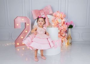 A toddler girl in a pink ruffled dress poses beside a lit number 2 at her birthday photoshoot with floral decor.