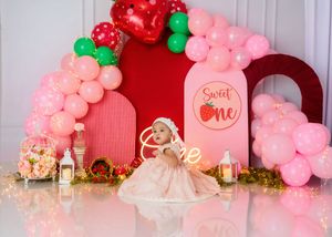 Baby in cream dress sits before Sweet One strawberry themed first birthday balloon backdrop.