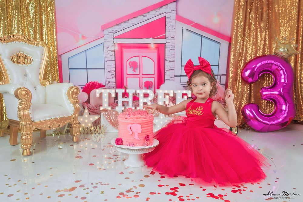 Toddler girl in red tutu dress poses with pink cake at Barbie-themed third birthday party celebration.