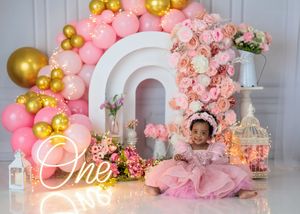 Baby girl in pink dress sits before white rainbow arch with pink roses, gold balloons, and One neon sign.