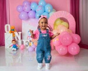 Toddler girl in denim overalls stands before colorful balloon arch with 'ONE' letters and toy decorations.