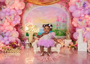 Toddler girl in pink tutu dress stands before princess castle backdrop with balloons and flowers for birthday.