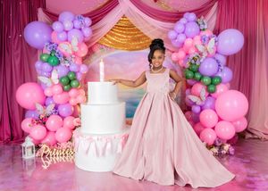 A young girl in a pink ballgown poses beside a tall white birthday cake with balloon arch and pink backdrop.