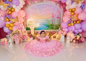 Baby in pink princess gown sits before castle backdrop with pink and purple balloons for first birthday.