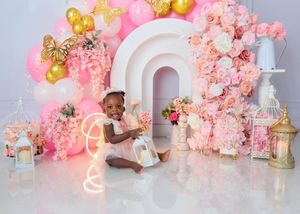 Baby girl in white outfit sits among lanterns and pink floral arch birthday photo setup.