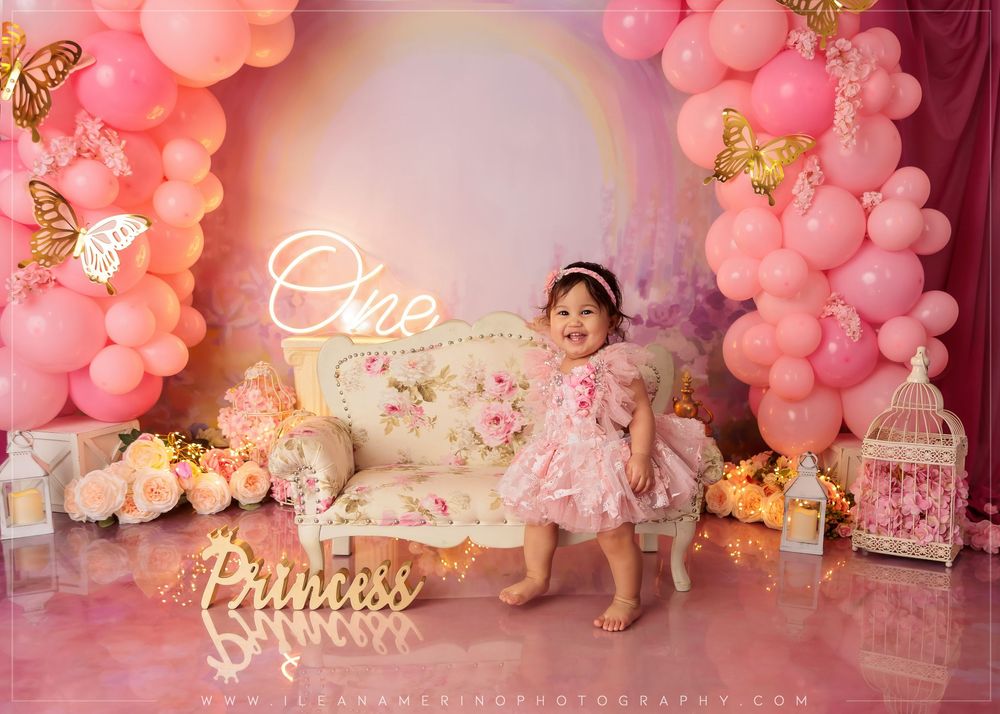 Baby girl in pink dress stands by floral sofa at butterfly princess first birthday party with pink balloons.