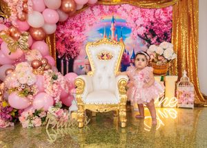 Baby girl in pink dress sits on white throne chair at princess first birthday party setup.
