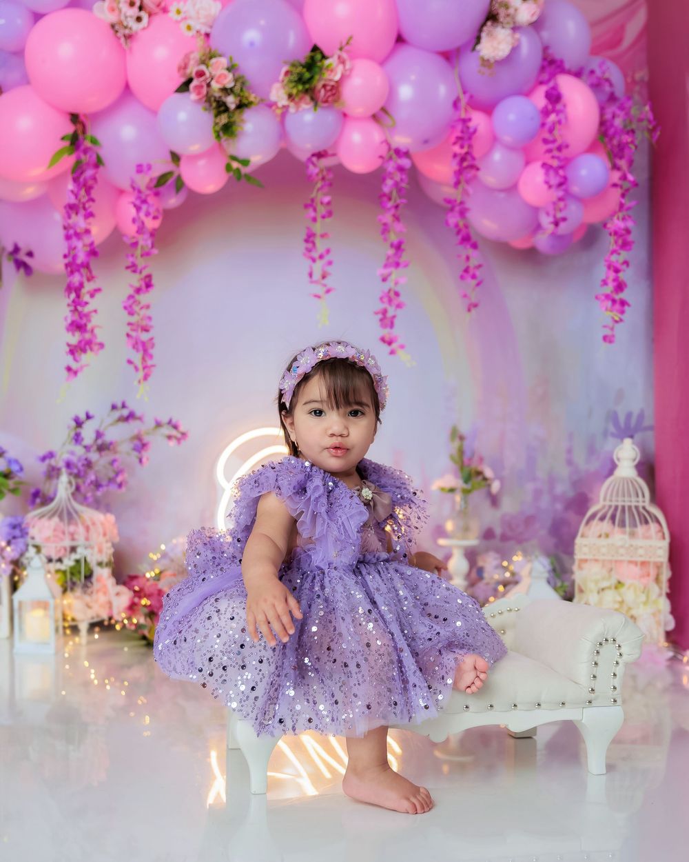 Baby girl in purple sequin dress stands at floral purple balloon arch birthday party with birdcage decor.