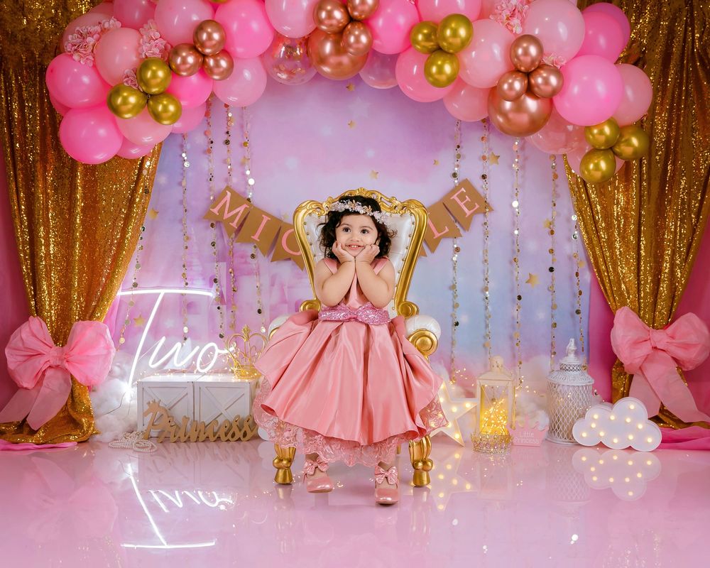 A toddler girl in a pink dress sits on a gold throne chair surrounded by pink and gold balloon arch decorations.