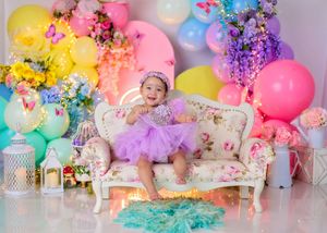 Happy baby girl in purple tutu sits on floral sofa surrounded by colorful balloons and flowers.