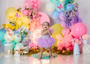 Toddler girl in purple tutu dress stands clapping before colorful balloon arch with flowers and fairy lights.