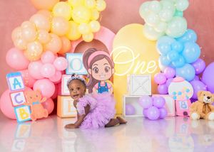 A baby girl in a purple tutu dress poses at her first birthday photoshoot with colorful balloons and alphabet blocks.