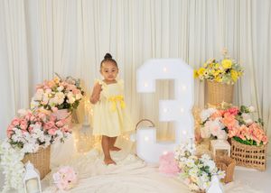 A toddler girl in a yellow dress stands beside a lit number 3 at her birthday photoshoot with floral arrangements.
