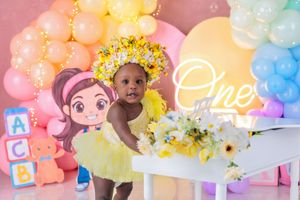 A baby girl in a yellow tutu dress stands at a white piano at her first birthday photoshoot with balloon backdrop.