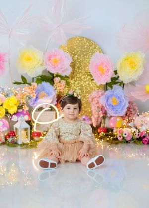 Smiling baby girl in pink tutu sits before large paper flowers and gold sequin backdrop with fairy lights.