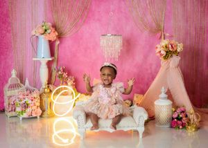 Smiling baby girl in pink dress sits on white chaise with chandelier and floral decor on pink backdrop.