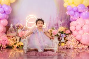 A baby girl in a white dress sits on a white chair surrounded by colorful balloons and flowers at her birthday shoot.