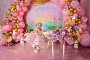 Baby girl in pink dress sits by floral table with pink gold balloon arch and butterfly decorations.