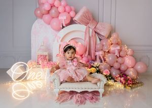 Baby girl in pink dress sits on white sofa surrounded by pink balloons and One neon sign for first birthday.