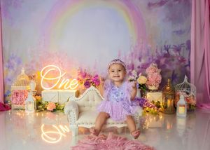 Baby in purple romper sits on white sofa with rainbow backdrop, birdcages, flowers, and One neon sign.