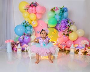Baby girl in purple tutu sits before rainbow balloon arch with flowers and lanterns on white backdrop.