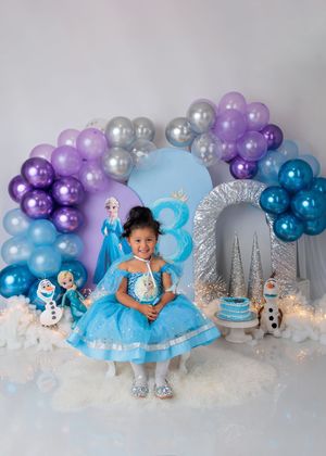 Young girl in blue Frozen Elsa dress poses before purple balloon arch with Elsa and Olaf figures for birthday.