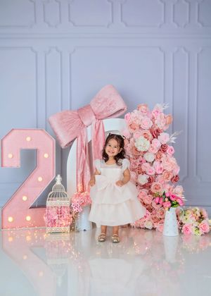 Toddler girl in white dress poses by illuminated number 2 and pink floral arrangement.