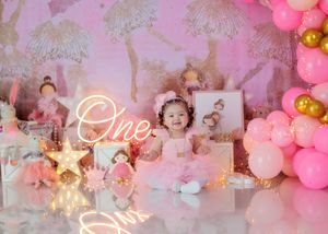 Baby girl in pink tutu sits at ballerina themed first birthday setup with One neon sign.