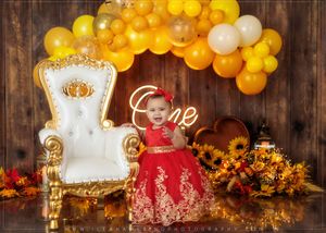 Baby girl in red dress sits before white throne chair with sunflowers, yellow balloons, and One neon sign.