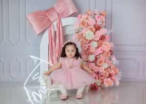 Toddler girl in pink dress poses beside floral number one and large pink bow decoration.