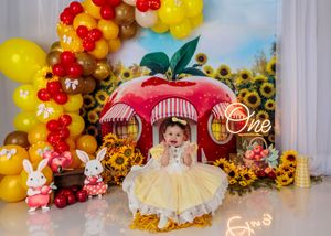 Baby in yellow dress sits before giant apple backdrop with sunflowers, bunnies, and balloons for first birthday.
