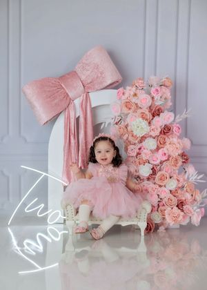Toddler girl in pink dress sits on white bench beside floral arrangement with Two neon sign.
