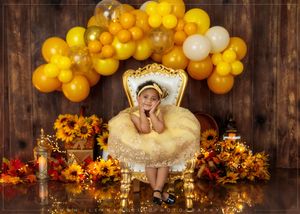 Smiling toddler girl in yellow dress sits on golden chair surrounded by sunflowers and yellow balloons.