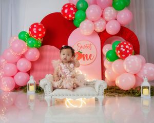 Baby girl in pink dress sits on white sofa at strawberry-themed first birthday cake smash with balloon arch.