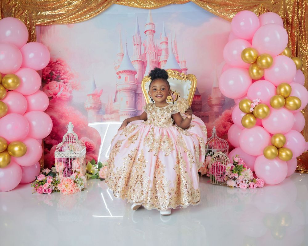 Little girl in gold embroidered gown sits on throne at pink castle-themed birthday party with balloon columns.