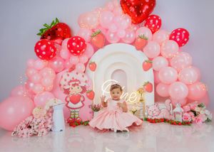 Baby girl in pink dress sits before strawberry-themed balloon arch with neon name sign backdrop.