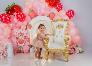 Smiling baby girl in pink dress stands beside ornate gold chair with strawberry balloon arch backdrop.