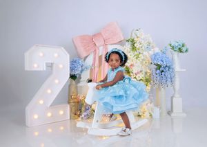 Toddler girl in blue ruffled dress sits on rocking horse beside illuminated number 2 with flowers.